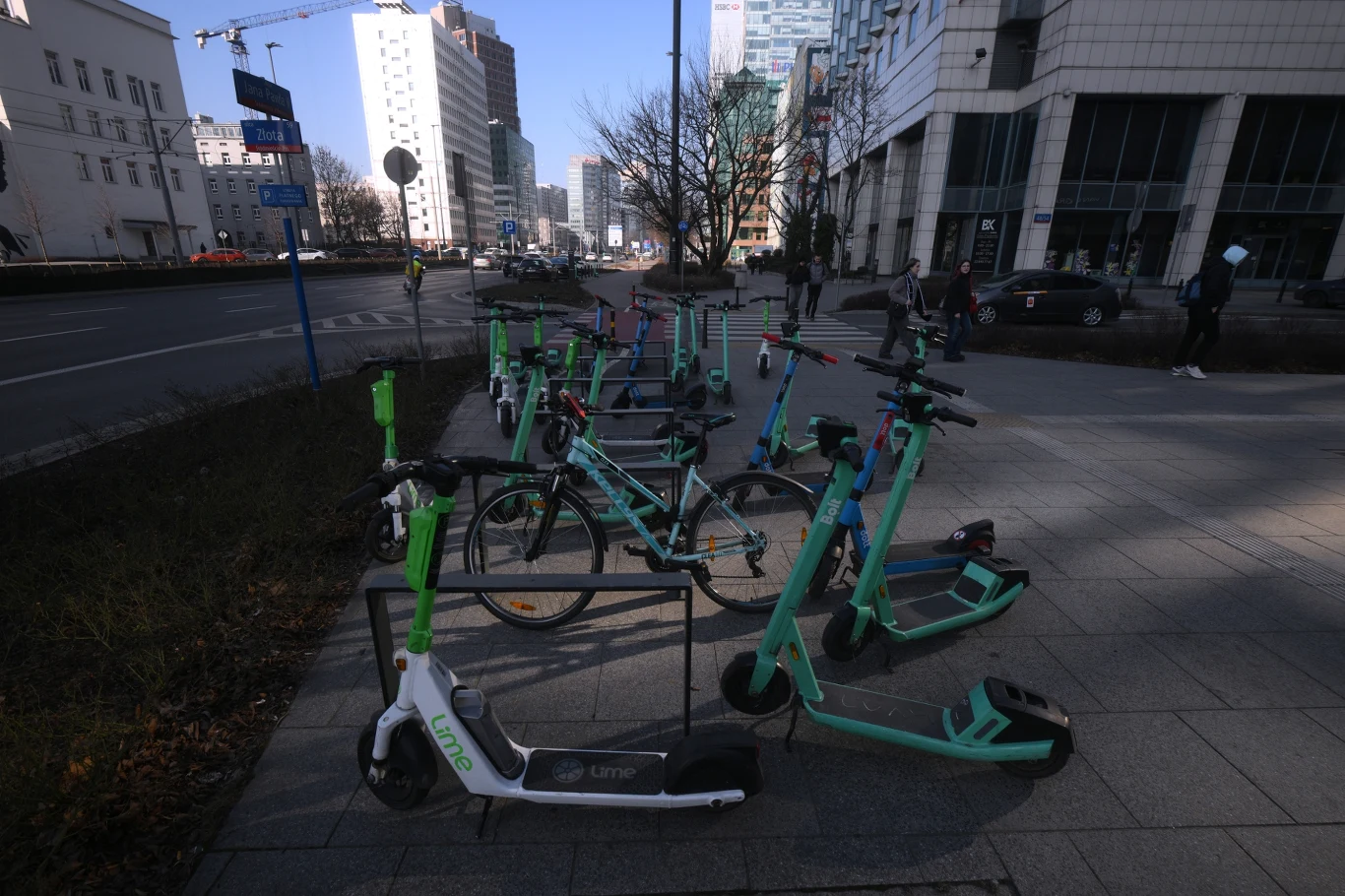 Policja wystawiła ponad 100 mandatów użytkownikom hulajnóg Rząd elektrycznych hulajnóg i rowerów miejskich ustawionych na chodniku wzdłuż ulicy w centrum miasta, w tle nowoczesne budynki biurowe i kilka przechodzących osób.