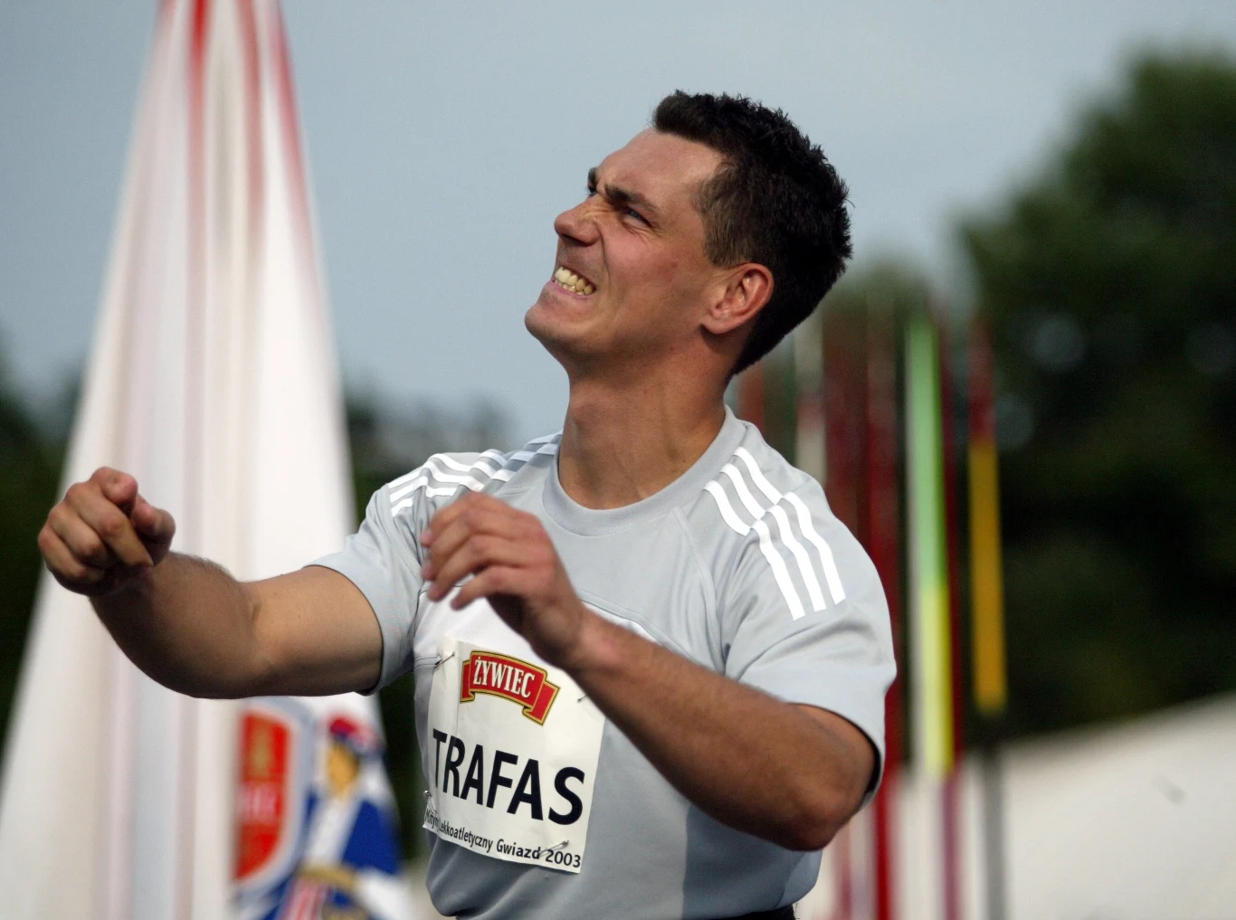 Dariusz Trafas Atleta w sportowej koszulce z nazwiskiem 'Trafas' wyraża intensywne emocje podczas zawodów lekkoatletycznych, w tle widoczne maszty i rozmyte elementy stadionu.