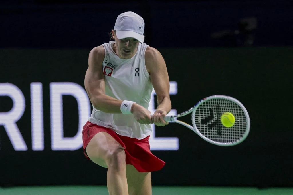 Tenisistka uderza piłkę podczas meczu tenisowego, ubrana w sportowy strój z białą czapką i czerwoną spódniczką, dynamiczna akcja na tle czarnej tablicy z napisem.