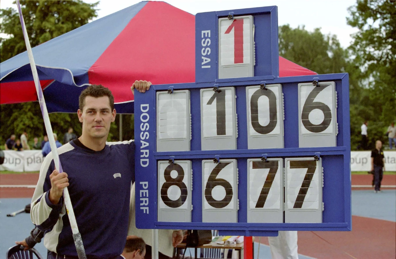Dariusz Trafas Mężczyzna stojący obok tablicy wyników lekkoatletycznych, trzymający tyczkę, z dużymi cyframi wskazującymi osiągnięty wynik w zawodach sportowych. W tle widoczne elementy stadionu lekkoatletycznego i kolorowy namiot.