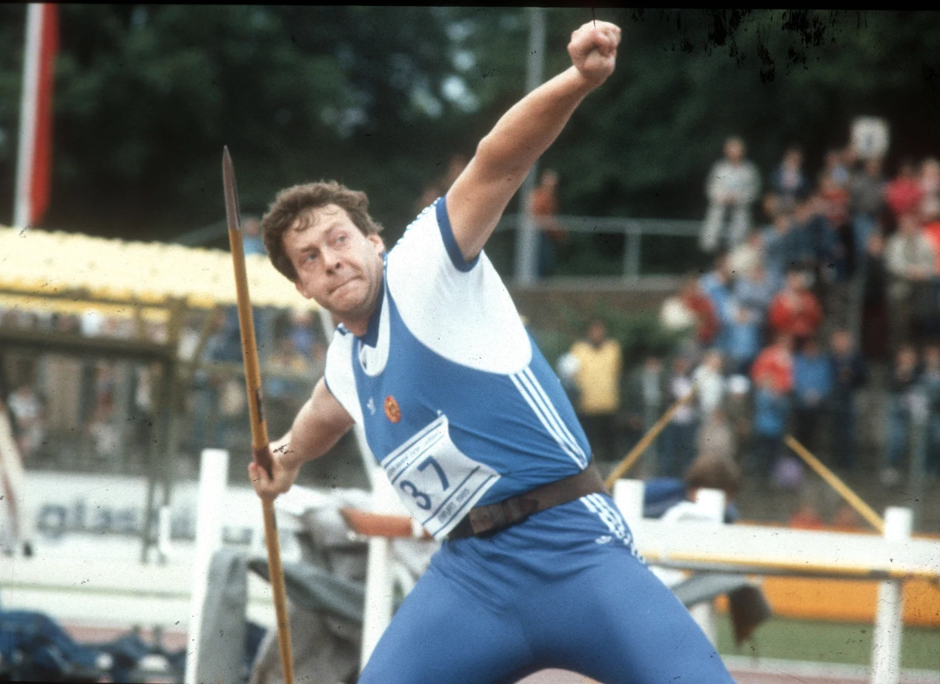 Uwe Hohn Sportowiec w niebieskim stroju wykonuje rzut oszczepem na stadionie lekkoatletycznym podczas zawodów, w tle widoczni są kibice na trybunach.