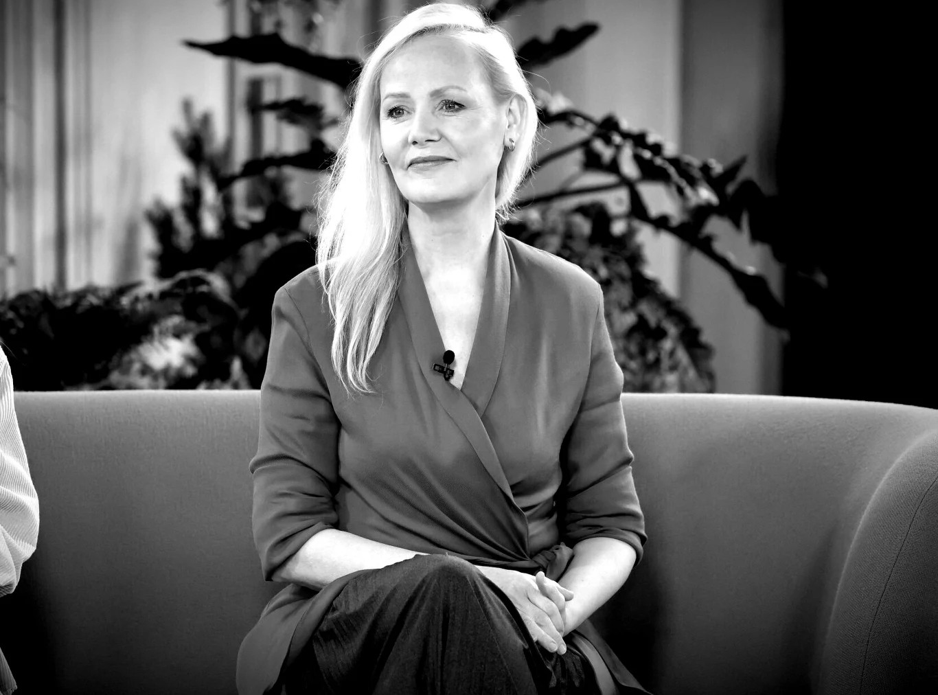 Katarzyna Stoparczyk nie żyje Kobieta z długimi blond włosami siedzi na kanapie, ubrana w elegancką sukienkę z długim rękawem, z zapiętym przypiętym mikrofonem, w tle widoczne rośliny.