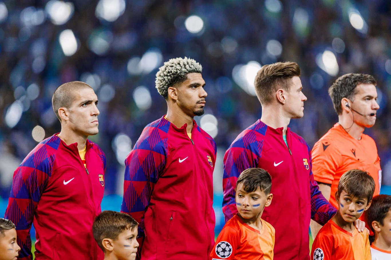 Oriol Romeu, Ronald Araujo i Marc Andre ter Stegen Piłkarze w czerwonych bluzach klubowych stoją w szeregu na stadionie przed rozpoczęciem meczu, obok nich znajdują się dzieci w pomarańczowych koszulkach oraz sędzia w pomarańczowej koszulce. W tle zamazane światła i publiczność.