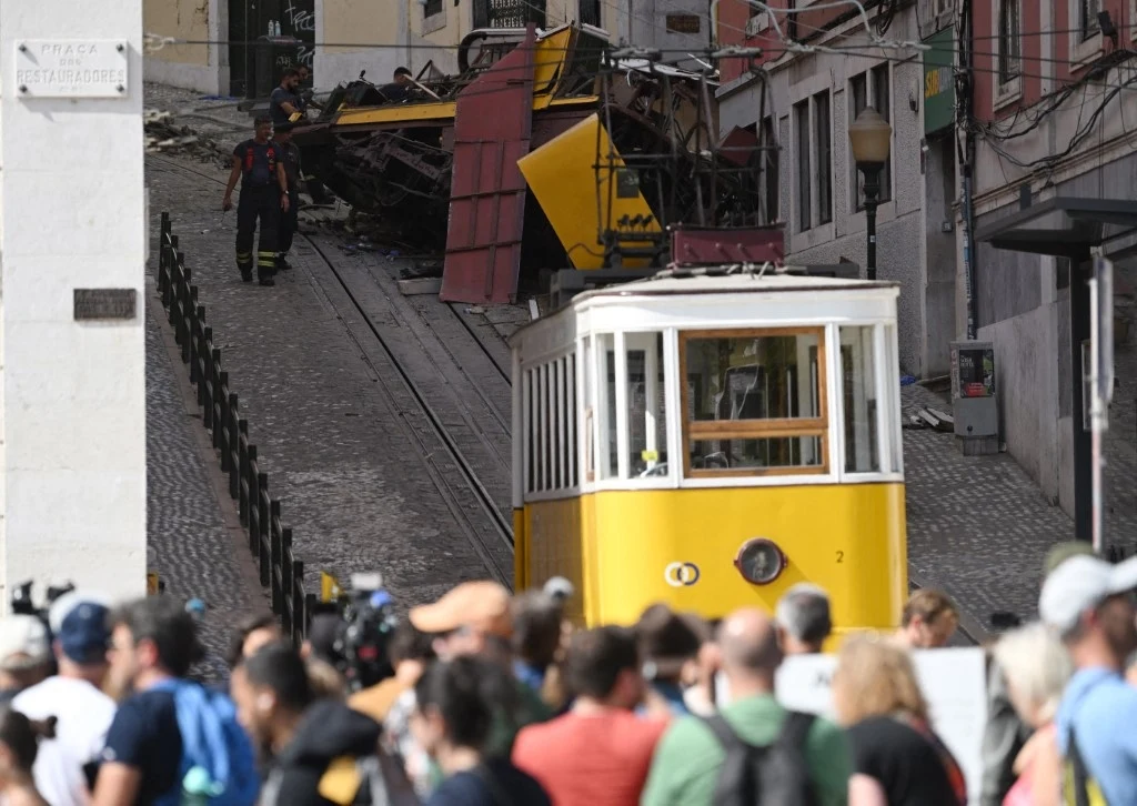 Tragiczny wypadek kolejki w Lizbonie Tłum ludzi zgromadzony na ulicy obserwuje miejsce wypadku tramwajowego, na pierwszym planie żółty tramwaj, w tle zniszczony wagon oraz służby ratunkowe.