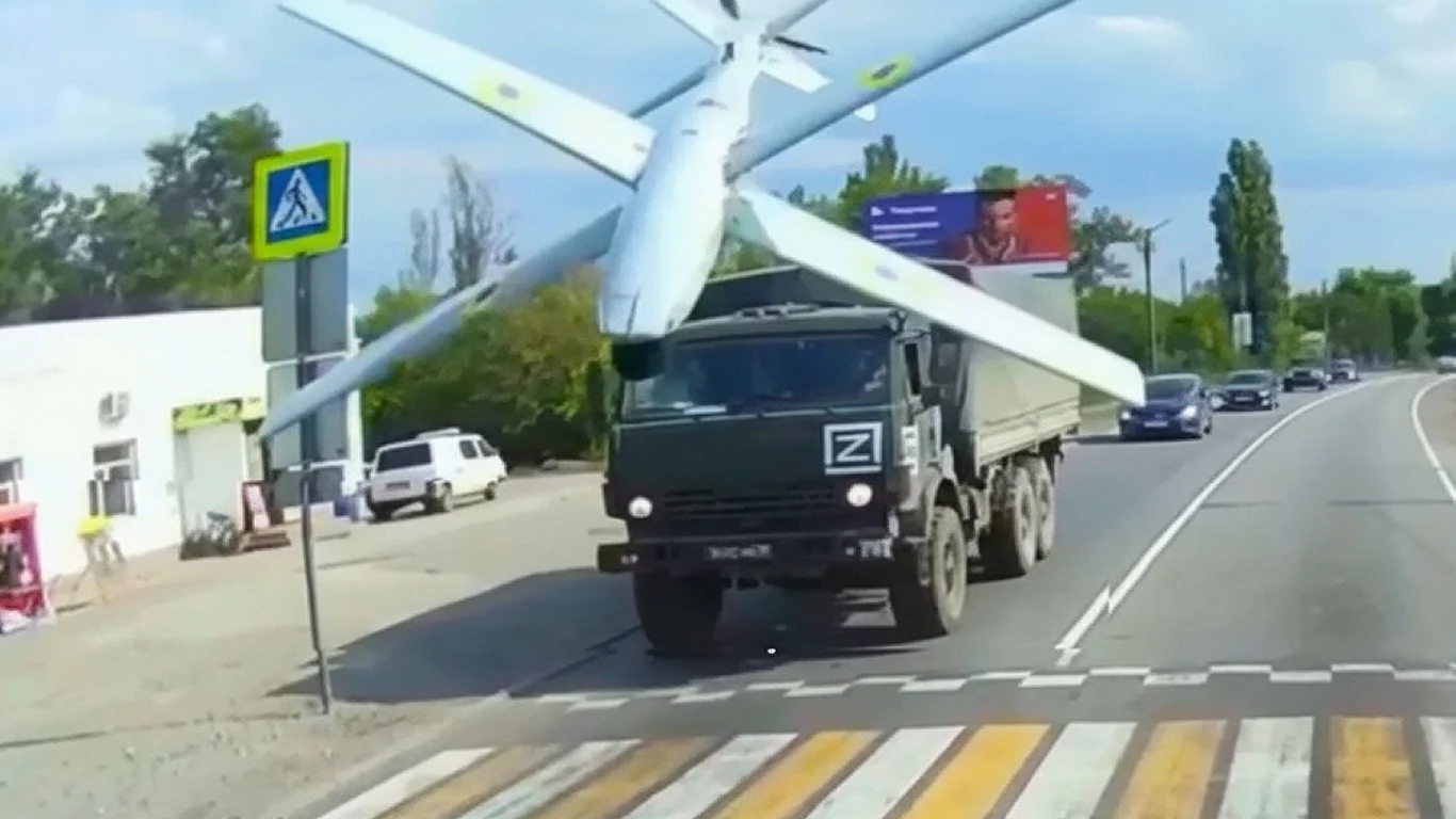 Niezwykłe nagranie uderzenia tajnego drona RAM-2X w rosyjski pojazd Duży wojskowy samochód ciężarowy z literą Z na masce transportuje bezzałogowy samolot wojskowy na otwartej drodze, w tle widzimy kilka samochodów, budynki oraz znaki drogowe.
