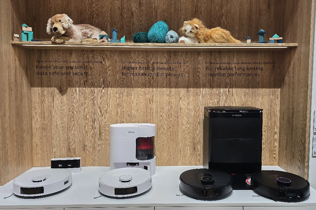 Nowoczesne roboty sprzątające różnych marek ustawione na białym blacie, w tle drewniana półka z pluszowymi kotami, motkami wełny i drobnymi dekoracjami, na ścianie widoczne anglojęzyczne opisy technologicznych zalet urządzeń.