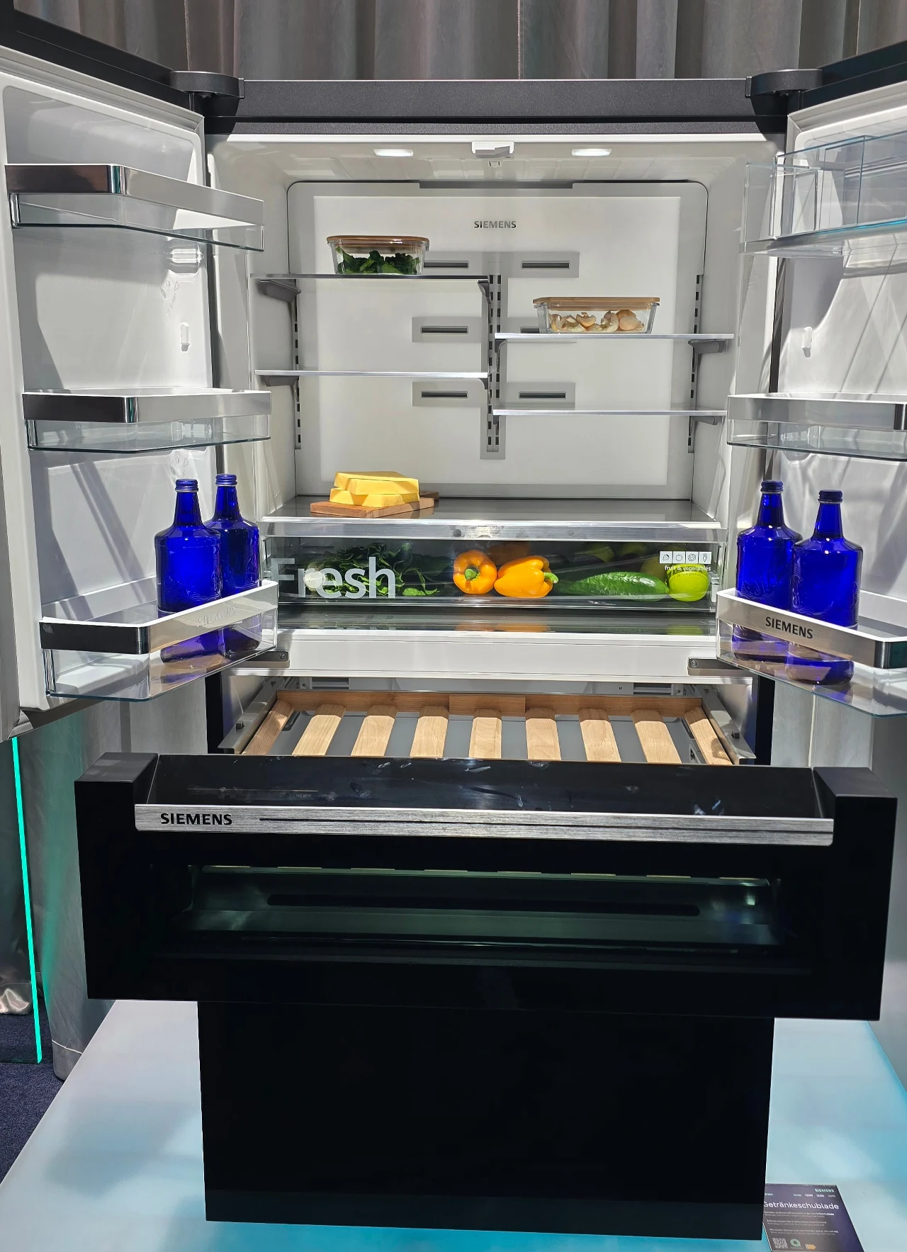 Nowoczesna lodówka marki Siemens z otwartymi drzwiami, w środku widoczne są przechowywane produkty spożywcze, takie jak warzywa, cytrusy, żółty ser, niebieskie butelki oraz kilka pojemników z jedzeniem. Wnętrze wyróżnia się czystością i ergonomicznym u...