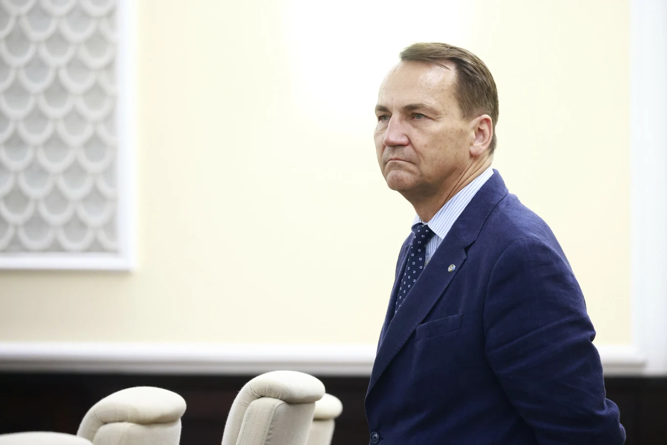 Wicepremier Radosław Sikorski zapytał Elona Muska o Starlinki, wykorzystywane przez Rosję do ataków dronowych w Ukrainie. Mężczyzna w granatowej marynarce i niebieskiej koszuli z krawatem w kropki stoi w jasnym, formalnym pomieszczeniu, na tle beżowej ściany i pustych krzeseł.