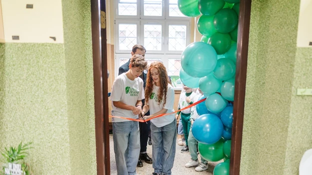 Dwie młode osoby przecinają czerwoną wstęgę podczas oficjalnego otwarcia, w otoczeniu dużego łuku z niebieskich i zielonych balonów, w tle widoczne są inne osoby oraz duże okno.
