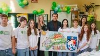 Grupa młodych osób i dwoje dorosłych stoi razem w jasnym pomieszczeniu, trzymając duży plakat z kolorową ilustracją przedstawiającą miasto Jawor oraz herb miasta, wszyscy noszą białe koszulki z zielonym logo Czyste Zielone Miasta, w tle balony i dekora...