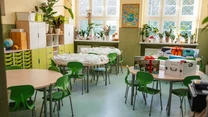 Kolorowa sala lekcyjna z okrągłymi stołami i zielonymi krzesłami, liczne rośliny doniczkowe na parapetach i szafkach, na stołach leżą różne przybory oraz pudła z materiałami edukacyjnymi. Jasne światło wpada przez okna, tworząc przyjazną atmosferę.