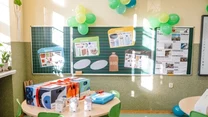 Sala szkolna udekorowana zielonymi i żółtymi balonami, z okrągłym stołem, na którym znajdują się zapakowane prezenty oraz materiały edukacyjne do recyklingu. Tablica ozdobiona jest plakatami na temat segregacji odpadów, co sugeruje tematykę ekologiczną...