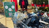 Kosz z dużą ilością zużytego sprzętu elektronicznego, takiego jak kable, suszarki, klawiatury i ładowarki, ustawiony na zewnątrz w pobliżu choinek i tablicy informacyjnej zachęcającej do recyklingu elektroniki.