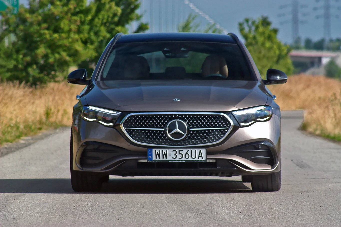 Szeroki przód luksusowego samochodu marki Mercedes-Benz z charakterystycznym logo na grillu, widoczny na pustej drodze otoczonej suchą trawą; nowoczesny design reflektorów i elegancki, metaliczny lakier w brązowym odcieniu.