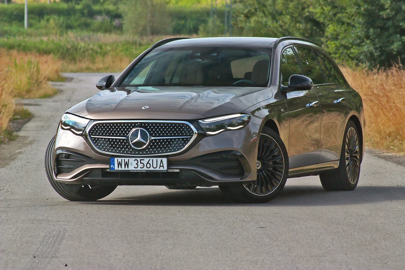 Nowoczesny samochód marki Mercedes klasy E w wersji kombi zaparkowany na bocznej, asfaltowej drodze otoczonej przez wysoką trawę i drzewa, dobrze widoczne przednie reflektory oraz charakterystyczna atrapa chłodnicy z logo producenta.