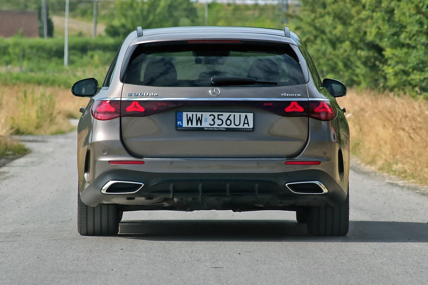 Tylna część srebrnego Mercedesa E 220d 4Matic kombi z polską tablicą rejestracyjną, jadącego wąską asfaltową drogą przez zielone tereny wiejskie.