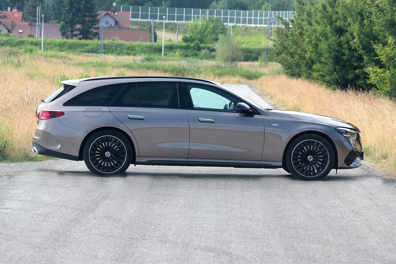 Srebrny samochód kombi Mercedes-Benz stoi na asfaltowej drodze, w tle widoczne są łąki, drzewa oraz zabudowania mieszkalne.