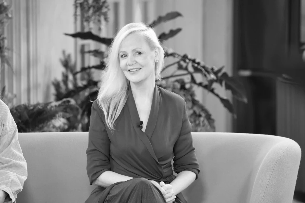 Uśmiechnięta kobieta o jasnych włosach siedzi na jasnej sofie w eleganckim wnętrzu, w tle widoczne są rośliny doniczkowe.