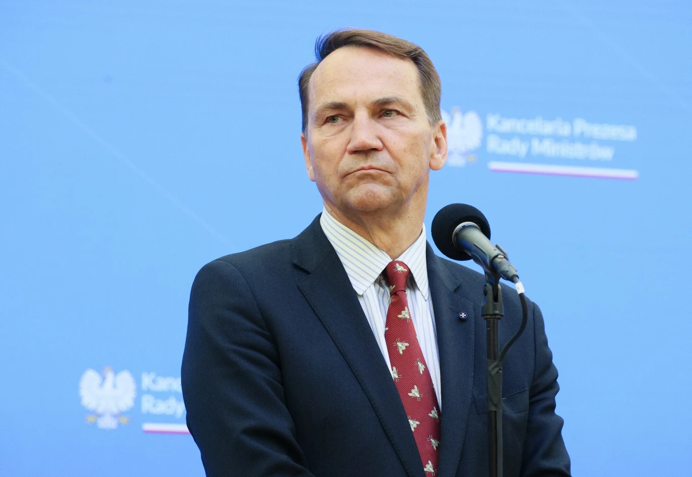 Radosław Sikorski odpowiedział polityko PiS Mężczyzna w garniturze i krawacie stoi przed mikrofonem na tle niebieskiej ściany z nadrukowanym napisem Kancelaria Prezesa Rady Ministrów oraz godłem Polski.