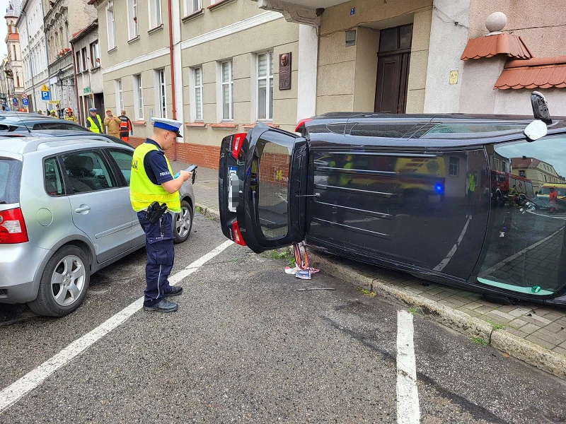 Policja poszukuje świadków wypadku w Chełmnie Wypadek samochodu osobowego przewróconego na bok na jezdni przy chodniku, policjant sporządzający notatkę, obok zaparkowany inny pojazd, w tle budynki mieszkalne oraz kilku obserwatorów.
