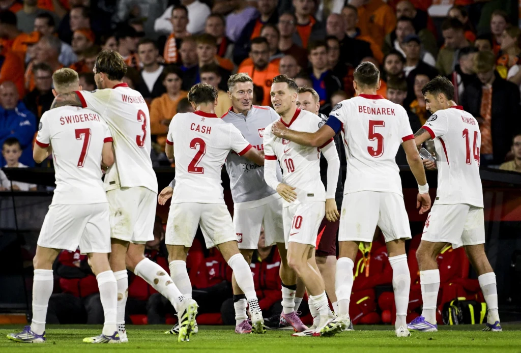 Piłkarze reprezentacji Polski w meczu z Holandią Grupa piłkarzy w białych strojach z czerwonymi akcentami cieszy się wspólnie ze zdobytej bramki podczas meczu, wokół nich widoczny stadion oraz kibice na trybunach.