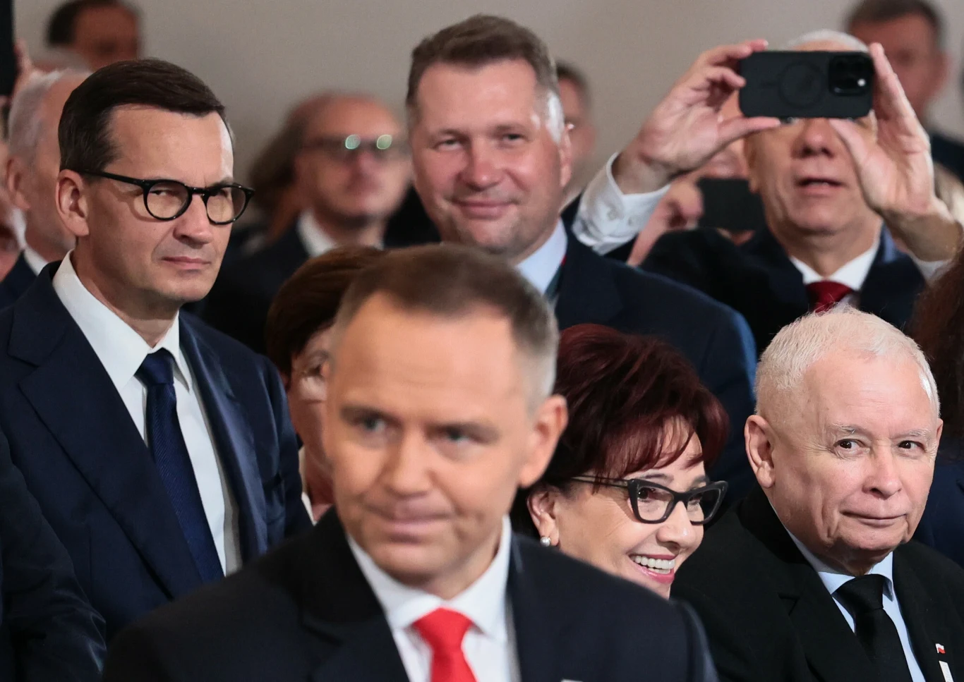 Jarosław Kaczyński nie kryje zadowolenia z przebiegu pierwszego miesiąca prezydentury Karola Nawrockiego Grupa polityków w garniturach siedzi podczas oficjalnego wydarzenia, jeden z mężczyzn robi zdjęcie telefonem, pozostali są skupieni lub uśmiechnięci.