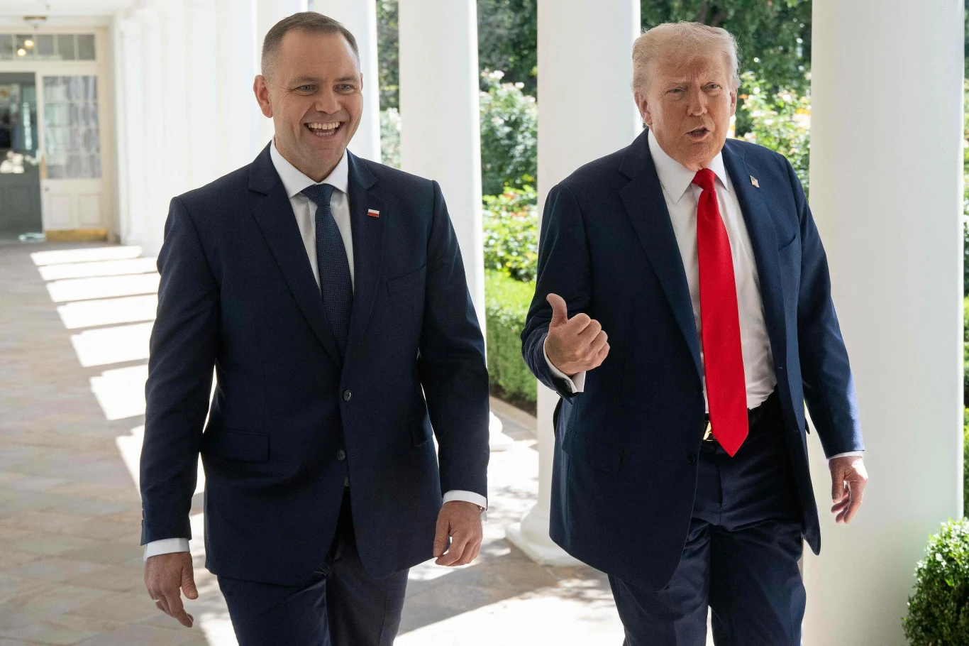 Prezydent Karol Nawrocki podczas spotkania z Donaldem Trumpem w Białym Domu Dwóch elegancko ubranych mężczyzn w garniturach idzie korytarzem na zewnątrz, jeden z nich energicznie gestykuluje, a drugi uśmiecha się, w tle kolumnada, zielone otoczenie i światło dzienne.