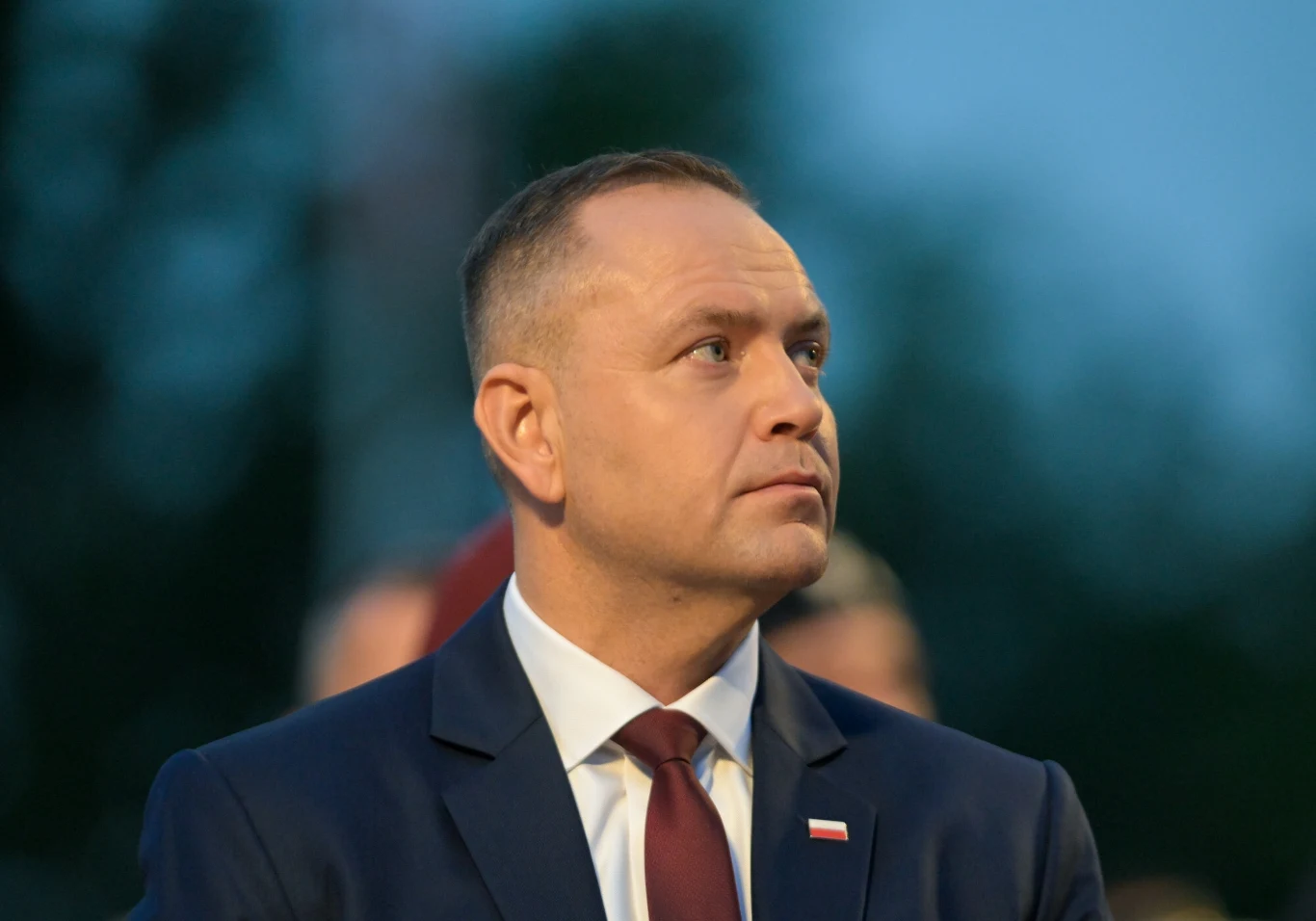 Prezydent Karol Nawrocki został zaprzysiężony 6 sierpnia 2025 roku Mężczyzna w garniturze i krawacie patrzący w bok, na klapie marynarki przypięta flaga Polski, rozmyte tło w odcieniach zieleni i niebieskiego.
