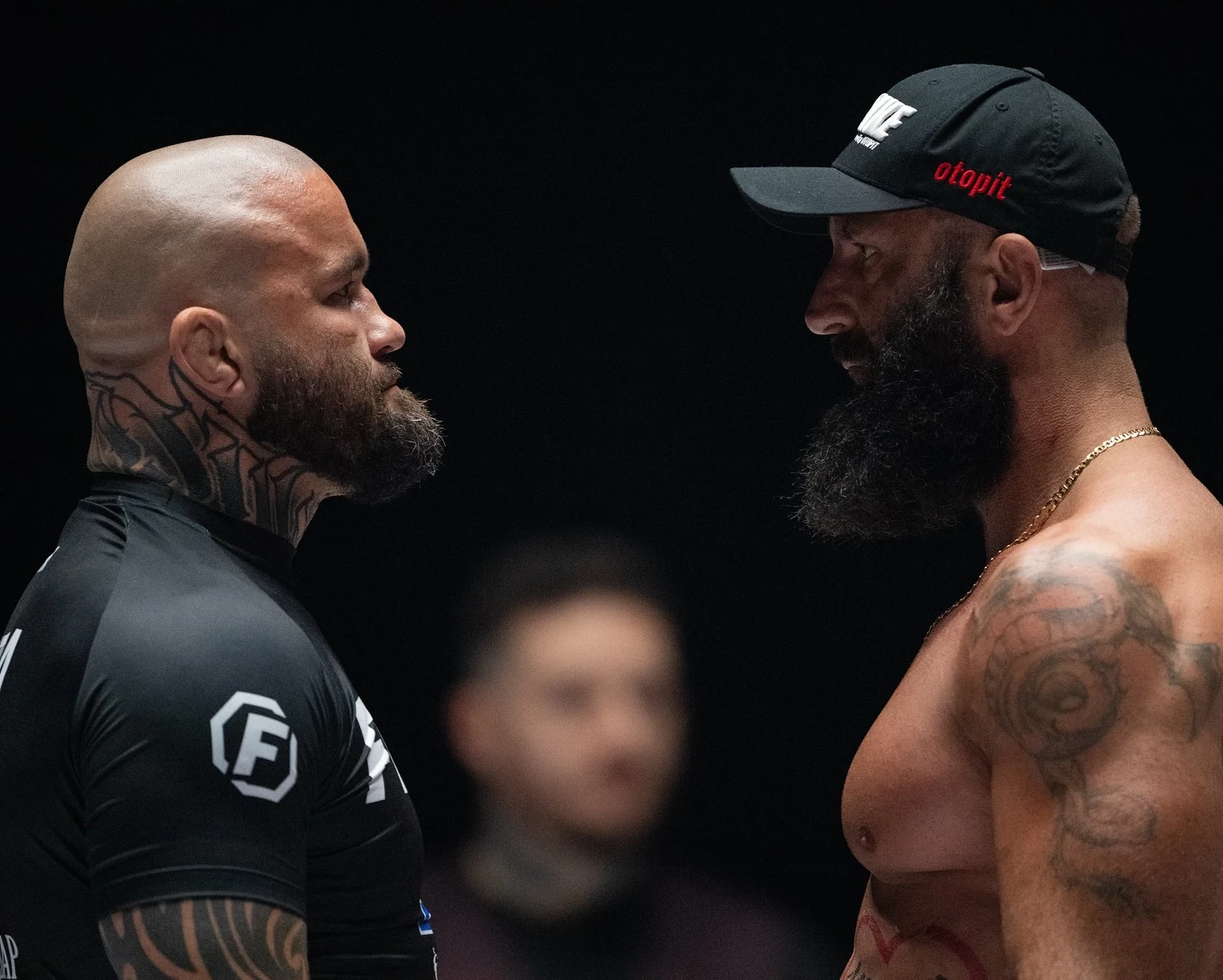 Dwóch umięśnionych mężczyzn mierzy się twarzą w twarz, jeden w sportowej koszulce i z tatuażami na głowie oraz rękach, drugi z brodą, goły od pasa w górę, w czapce i z widocznymi tatuażami, scena sugeruje moment napięcia przed walką sportową.