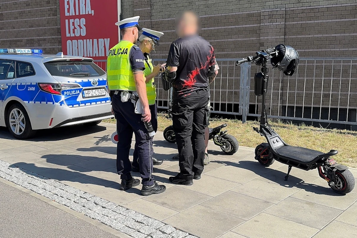 Policjanci kontrolowali użytkowników hulajnóg Dwóch policjantów w mundurach rozmawia z mężczyzną stojącym obok hulajnogi elektrycznej, obok policyjny radiowóz zaparkowany przy krawędzi chodnika oraz druga hulajnoga i kask przypięty do jej kierownicy.