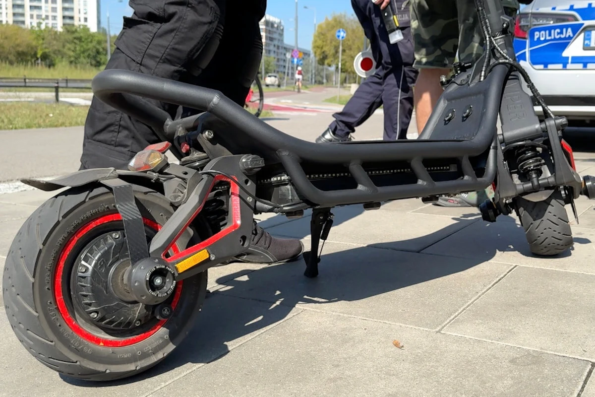 Prędkość maksymalna hulajnogi musi być ograniczona do 20 km/h Elektryczna hulajnoga o masywnej konstrukcji ustawiona na chodniku, w tle widoczne nogi kilku osób oraz fragment radiowozu policyjnego.