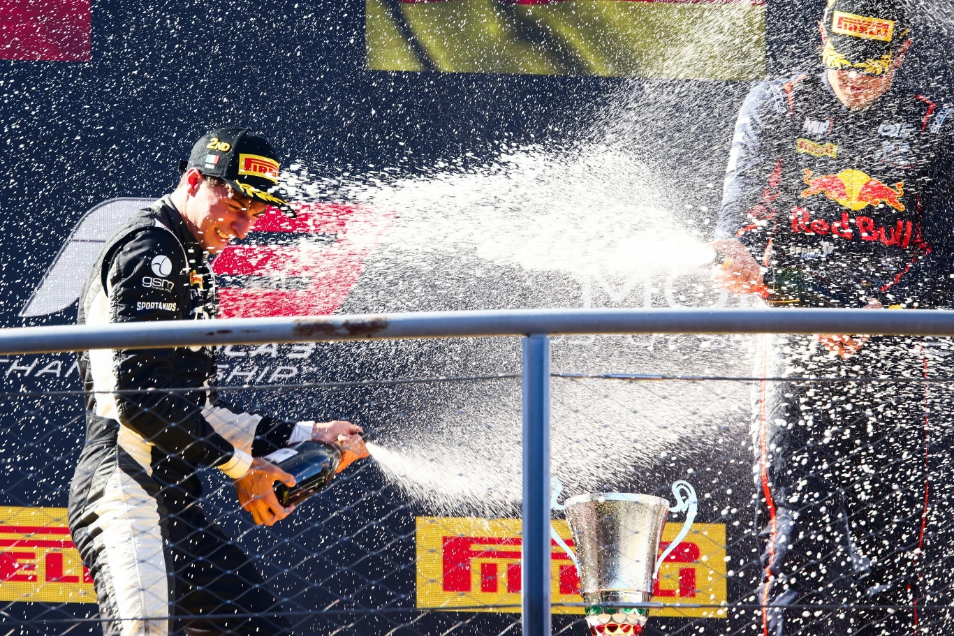 Dwóch kierowców wyścigowych w kombinezonach oraz czapkach z logo sponsorów świętuje na podium, rozpryskując szampana obok dużego pucharu i tablic sponsorskich.