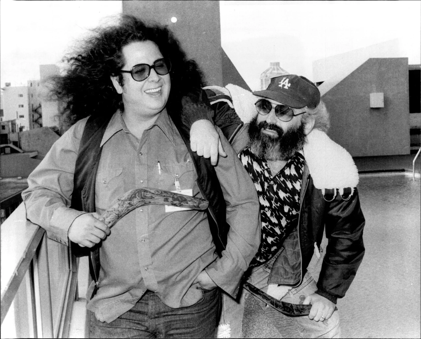 Mark Volman (Flo) i Howard Kaylan (Eddie) w 1976 r. Dwóch mężczyzn w okularach słonecznych, jeden z bujnymi kręconymi włosami i drugim w czapce z daszkiem oraz brodą, stoją obok siebie na zewnątrz, uśmiechają się i trzymają bumerangi, otoczona nowoczesną zabudową miejska sceneria