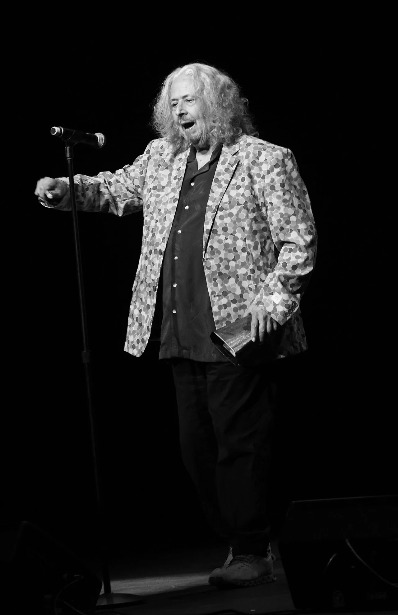 Mark Volman w czerwcu 2024 r. Mężczyzna z długimi, siwymi włosami w wzorzystej marynarce stoi na scenie przed mikrofonem i trzyma książkę, sprawiając wrażenie, że przemawia lub recytuje.