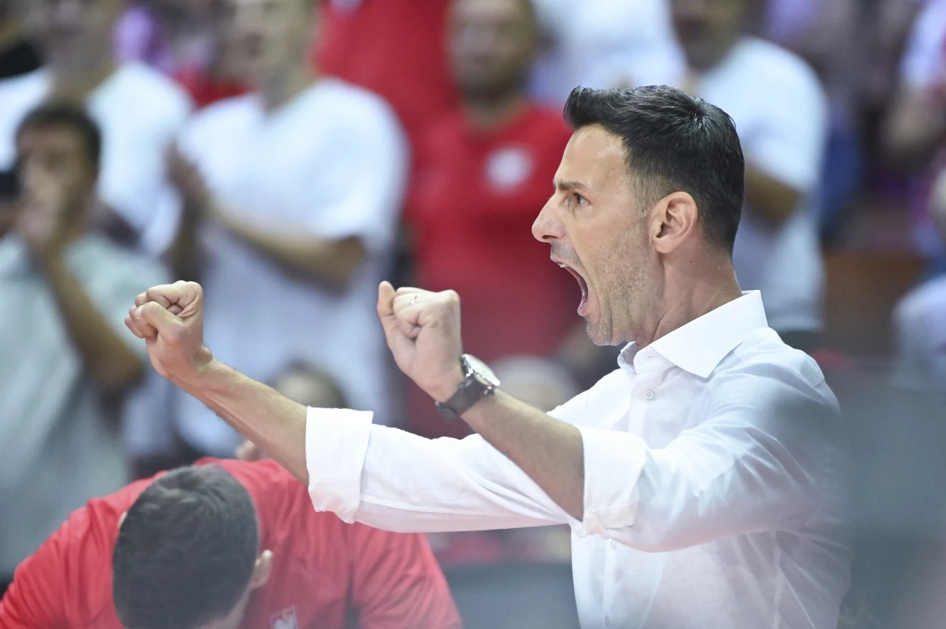 Mężczyzna w białej koszuli z uniesionymi pięściami wyraża silne emocje, prawdopodobnie radość lub entuzjazm podczas wydarzenia sportowego, w tle rozmyty tłum kibiców.