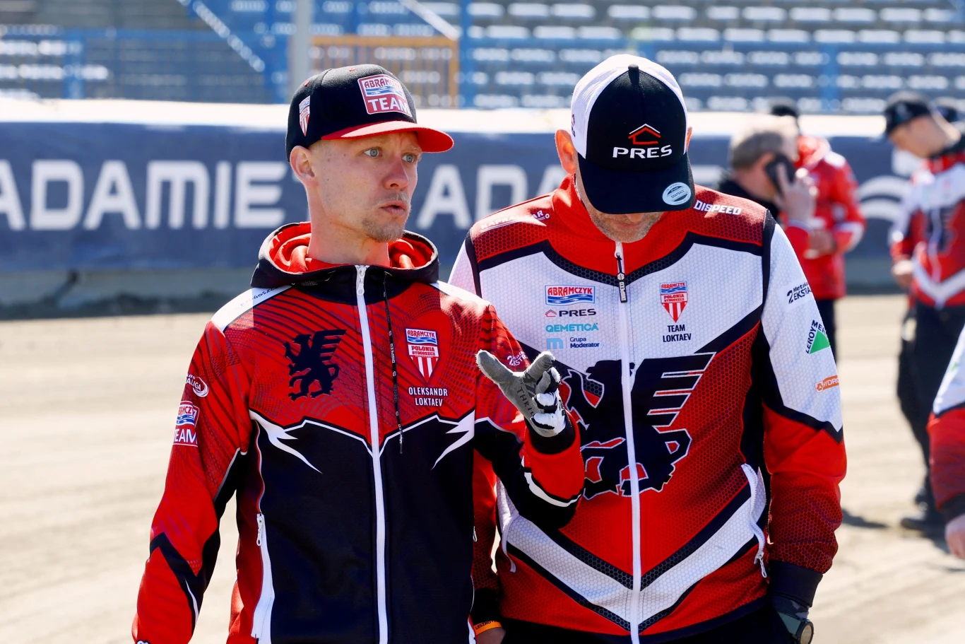 Aleksandr Łoktajew na obchodzie toru Dwóch mężczyzn ubranych w sportowe bluzy z logo drużynowym oraz czapki, rozmawia na stadionie żużlowym, w tle widoczni inni członkowie zespołu.