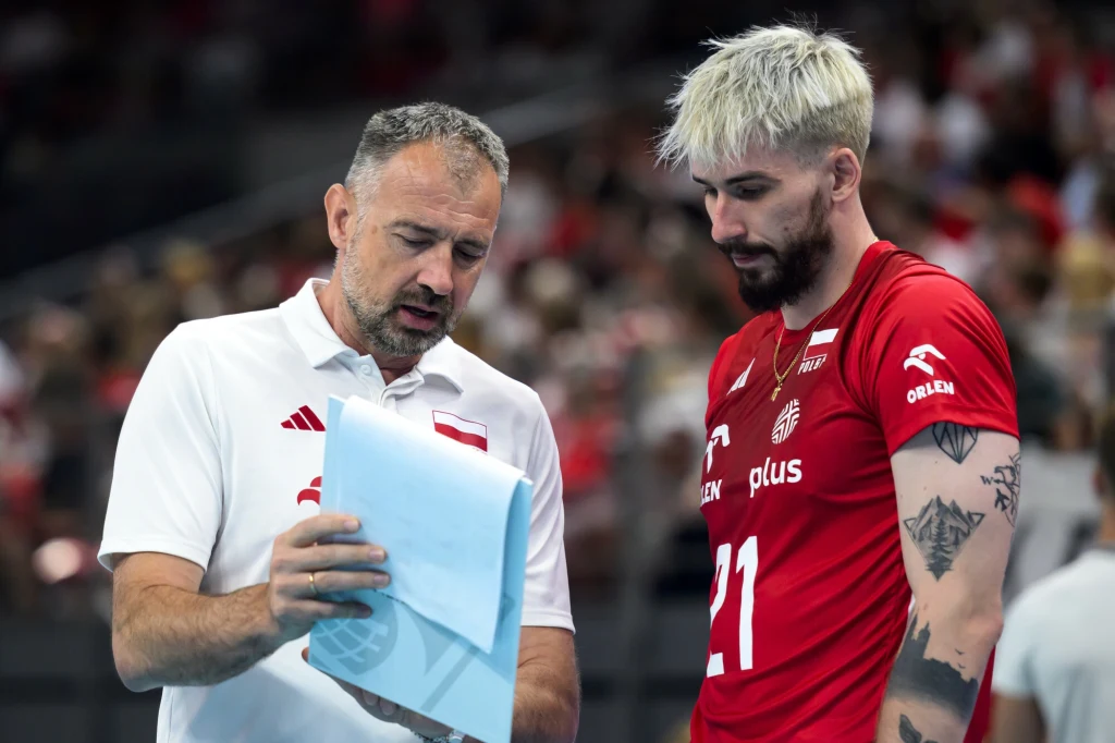 Nikola Grbić i Tomasz Fornal Trener w białej koszulce rozmawia z siatkarzem reprezentacji Polski w czerwonym stroju, pokazując mu notatki na kartkach. W tle widownia hali sportowej, atmosfera skupienia i koncentracji podczas rozgrywki.