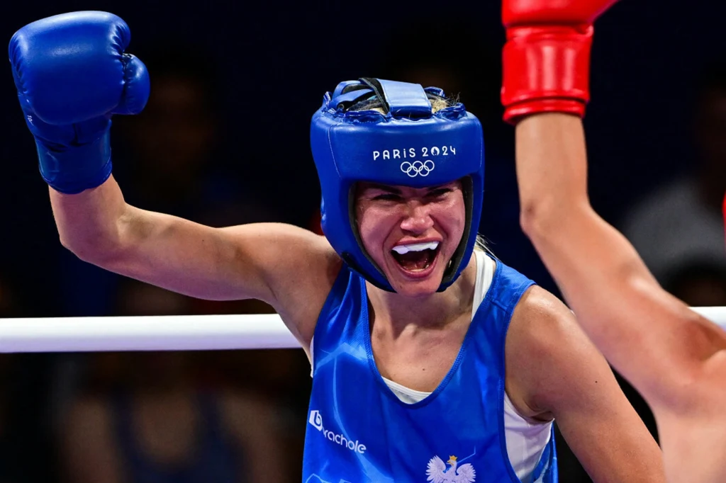 Aneta Rygielska Bokserka w niebieskim stroju z kaskiem ochronnym i rękawicami triumfalnie unosi rękę, na twarzy widać radość z sukcesu, w tle widoczny fragment ringu oraz rywal w czerwonych rękawicach.