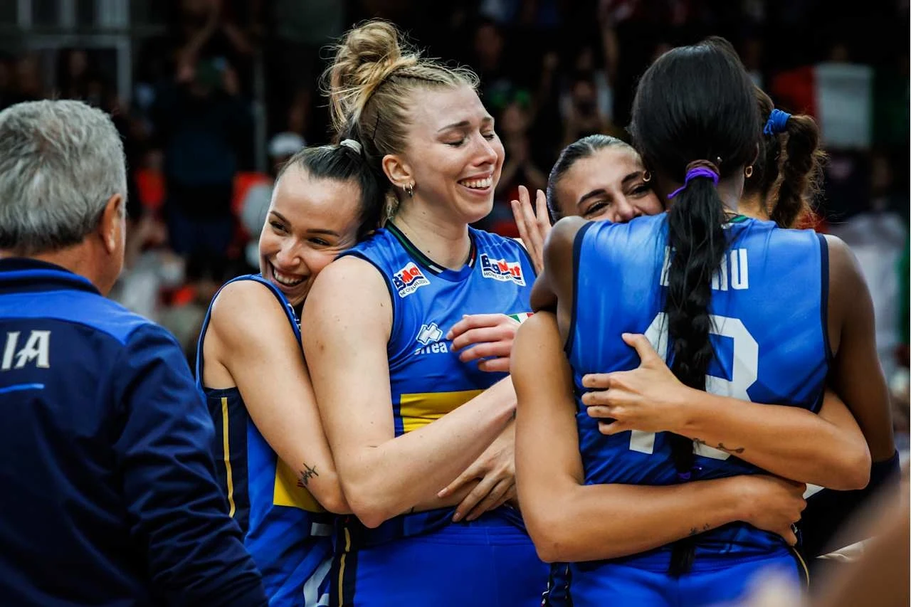 Siatkarki reprezentacji Włoch Grupa zawodniczek w niebiesko-żółtych strojach sportowych ściska się i uśmiecha, okazując radość i wzajemne wsparcie po zakończonym meczu siatkówki, a obok stoi trener w kurtce z napisem Italia.