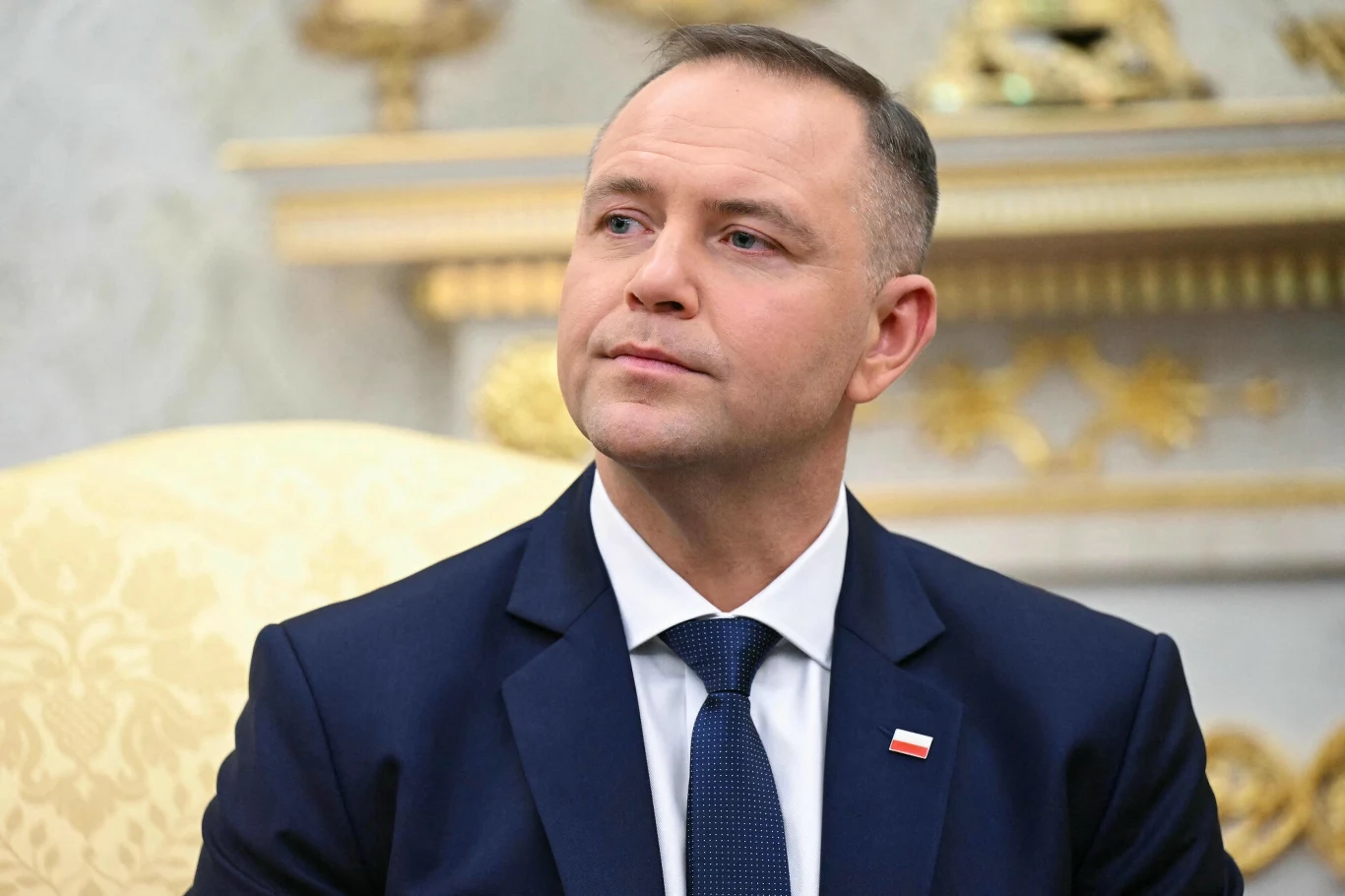 Prezydent Karol Nawrocki Mężczyzna w garniturze z przypiętą do klapy flagą Polski siedzi na jasnym fotelu, w tle zdobiona ściana i eleganckie elementy dekoracyjne.
