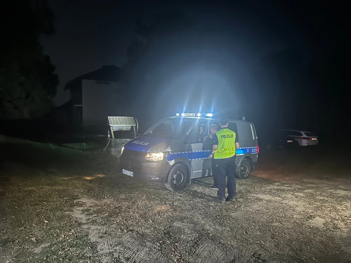 Majdan-Sielec. Niezidentyfikowany obiekt - prawdopodobnie dron - rozbił się na polu Policjant w odblaskowej kamizelce rozmawia przy radiowozie policyjnym zaparkowanym nocą na nieoświetlonym terenie, z tyłu widoczny jest budynek oraz inny samochód.