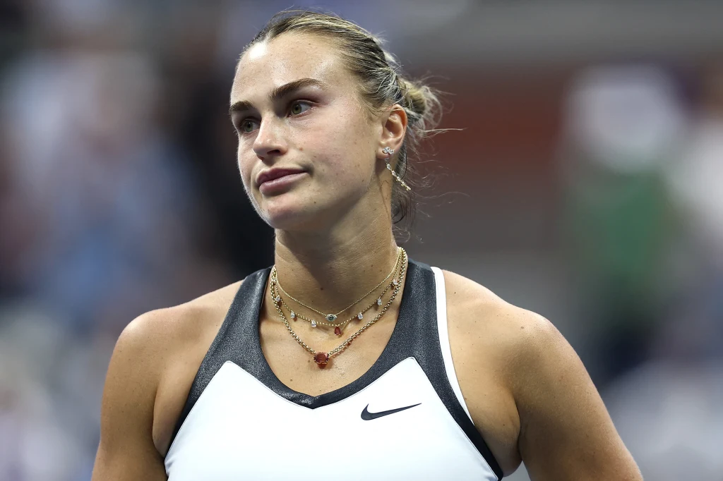 Aryna Sabalenka Kobieta w sportowym stroju tenisowym na korcie, wyraźnie skoncentrowana, nosi kilka delikatnych naszyjników, jej włosy są upięte, tło rozmyte.