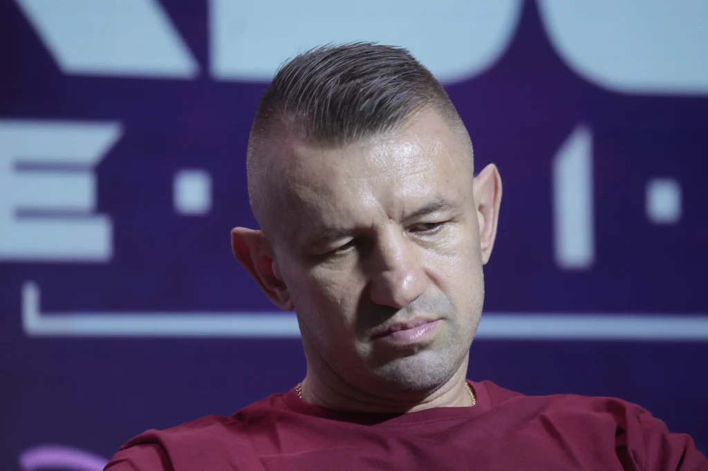 Mężczyzna w średnim wieku o krótkich, ciemnych włosach i opalonej cerze, ubrany w bordowy t-shirt, patrzy w dół z poważnym wyrazem twarzy na tle rozmytego, ciemnego napisu.