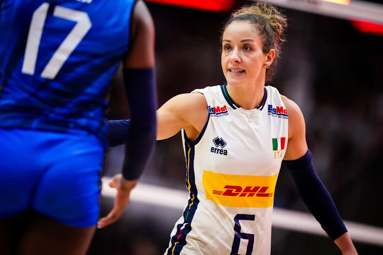 Monica De Gennaro, libero reprezentacji Włoch Siatkarka w białym stroju z numerem 6 i włoską flagą na piersi gestykuluje podczas meczu, naprzeciw niej widoczna jest zawodniczka w niebieskim stroju z numerem 17.