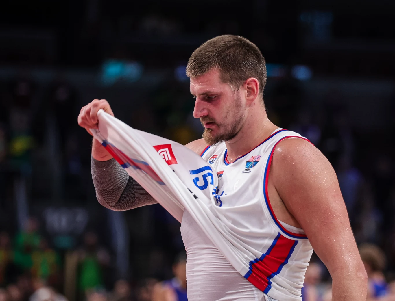 Nikola Jokić Koszykarz w białym stroju z numerem 15 unosi koszulkę do twarzy, sprawiając wrażenie skupienia lub zmęczenia podczas meczu sportowego.