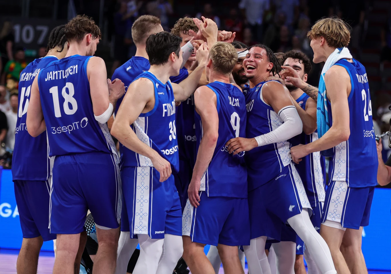 Finowie cieszą się z sensacyjnego awansu do ćwierćfinału EuroBasketu 2025 Grupa młodych koszykarzy w niebieskich strojach zgromadzona w kręgu na parkiecie, wyrażająca radość i ekscytację po wygranym meczu. W tle widać kibiców i jasne oświetlenie hali sportowej.
