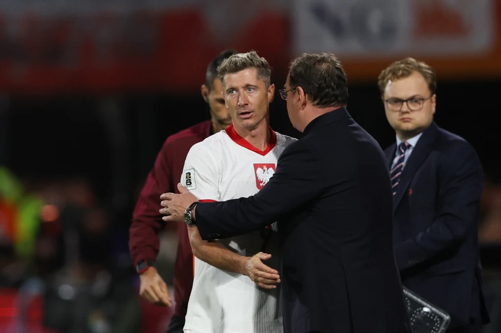 Robert Lewandowski i Jan Urban Piłkarz w białej koszulce z orłem na piersi stoi na boisku, otoczony przez mężczyzn w garniturach i sportowej odzieży; jedna z osób trzyma piłkarza za ramię.