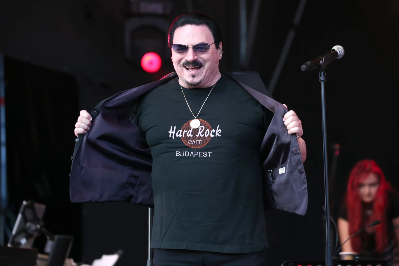 Bobby Kimball podczas Sziget Festival w 2017 r. Mężczyzna w okularach przeciwsłonecznych z szerokim uśmiechem rozchyla marynarkę, prezentując czarną koszulkę z logo Hard Rock Cafe Budapest, stojąc na scenie przy mikrofonie, w tle kobieta z czerwonymi włosami przy instrumentach.