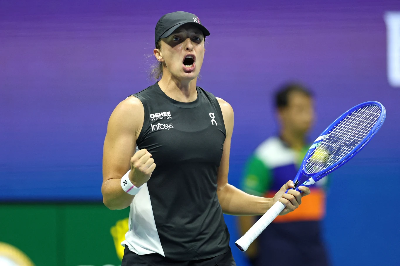 Iga Świątek Tenisistka w czarnym stroju sportowym trzyma rakietę tenisową i okazuje emocje, prawdopodobnie po udanym zagraniu, w tle rozmyta postać sędziego.