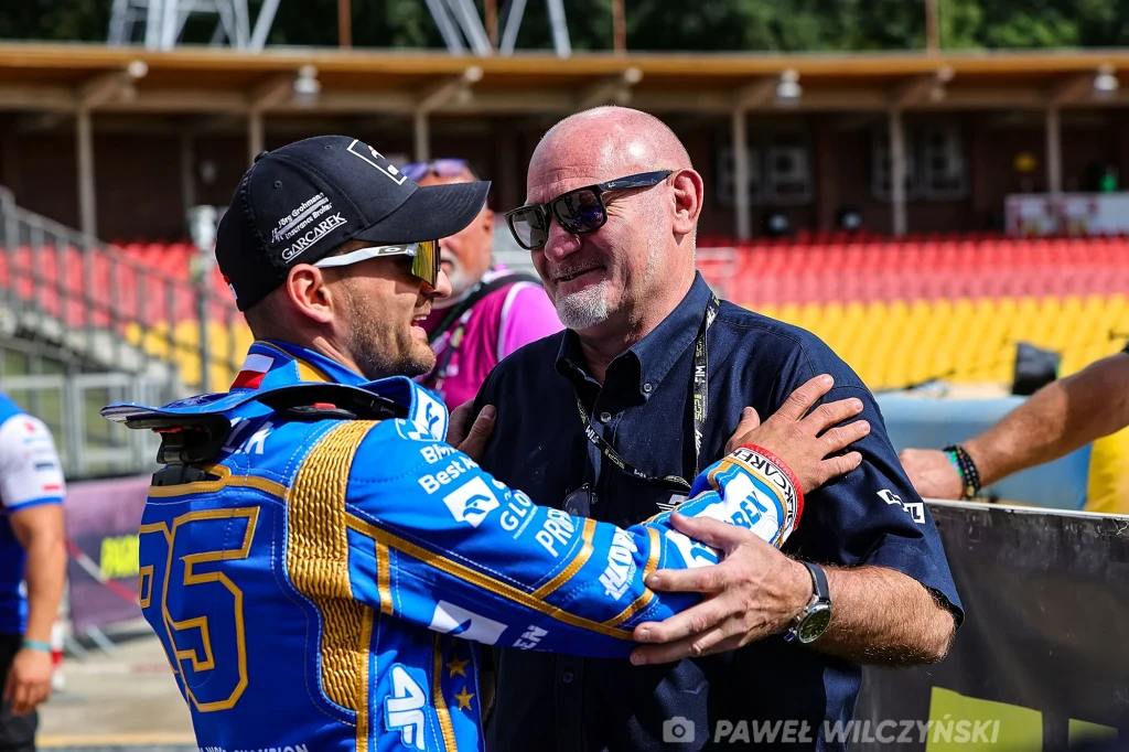 Dwóch mężczyzn obejmuje się i rozmawia na stadionie. Jeden z nich ubrany jest w sportowy kombinezon żużlowy z numerem 55, drugi ma na sobie ciemną koszulę i okulary przeciwsłoneczne.